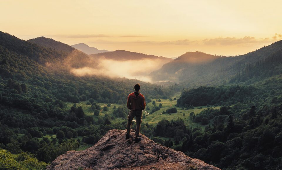 Young,Man,Standing,On,Top,Of,Cliff,In,Summer,Mountains